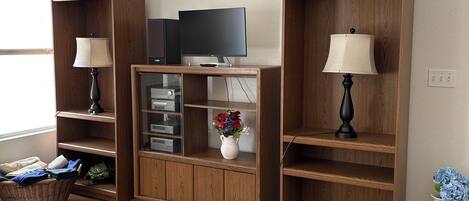 A living room with wooden shelving units, a flat-screen TV, a lamp, a vase with flowers, and a basket.