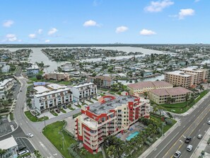 A coastal cityscape with a prominent hotel featuring balconies, a swimming pool, and a marina in the background.