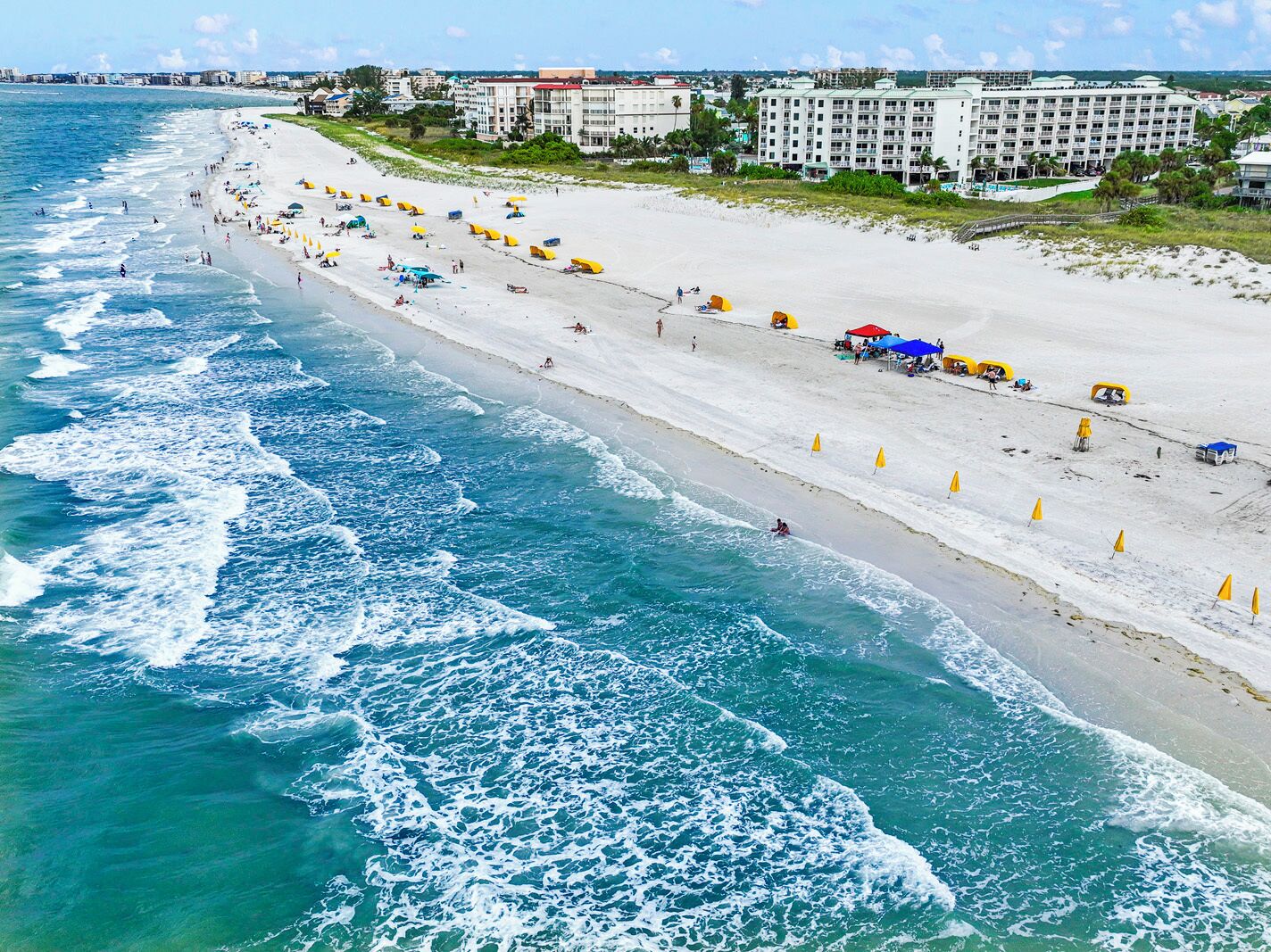 A beach with umbrellas and people, a hotel in the background, and waves crashing onto the shore.