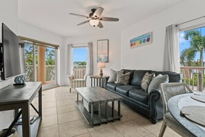 A spacious living area with a sofa, coffee table, and a television. There is a ceiling fan, a window with curtains, and a painting on the wall. - Sunset Views & Endless Balcony, On Site Heated Pool and Spa, Steps to Beach (Treasure Island)