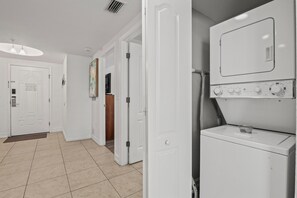A laundry room with a front-loading washer and dryer, a door leading to a hallway, and a light fixture on the ceiling. - Sunset Views & Endless Balcony, On Site Heated Pool and Spa, Steps to Beach (Treasure Island)