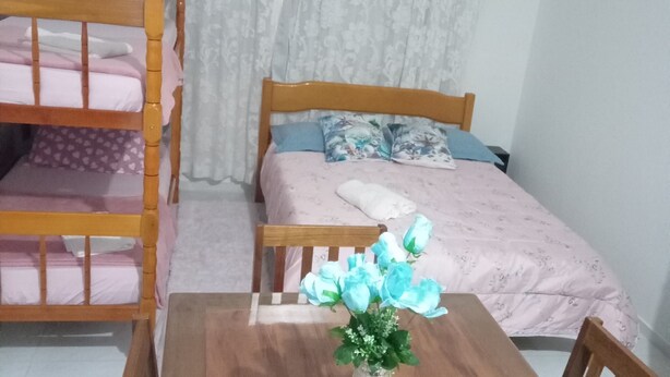 A room with a bunk bed, a single bed, a wooden table with chairs, and a vase with flowers. - Suite with kitchen, well located. (Barra Velha)