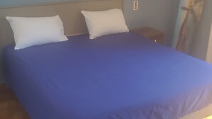 Un lit avec une couvre-lit bleue, deux oreillers blancs, une table de chevet, une lampe fixée au mur et un tableau accroché au mur.