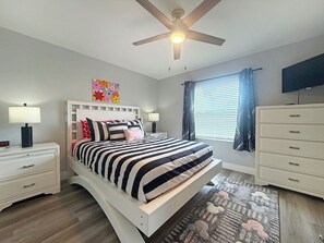 Un dormitorio con cama, mesitas de noche, ventilador de techo, ventana con cortinas, televisor y cómoda.