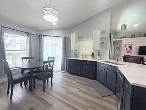 Una cocina moderna con gabinetes oscuros, una mesa de comedor con sillas, un ventanal con cortinas y un televisor montado en la pared.