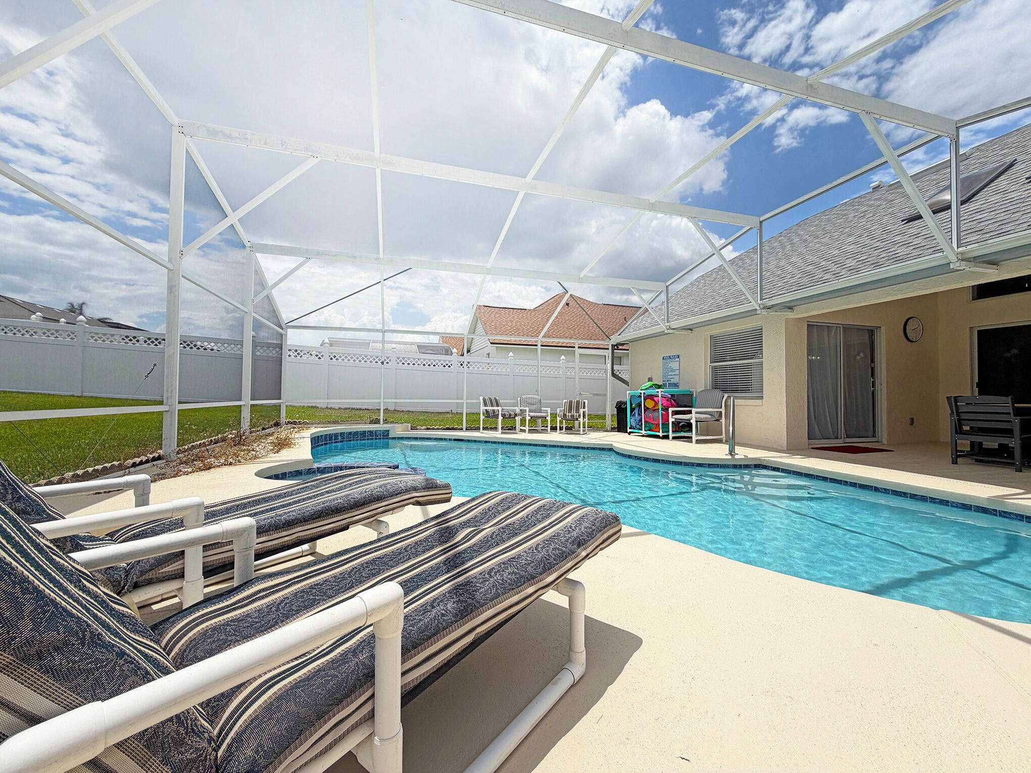A covered outdoor swimming pool with lounge chairs, a patio table, and a view of a house in the background.
