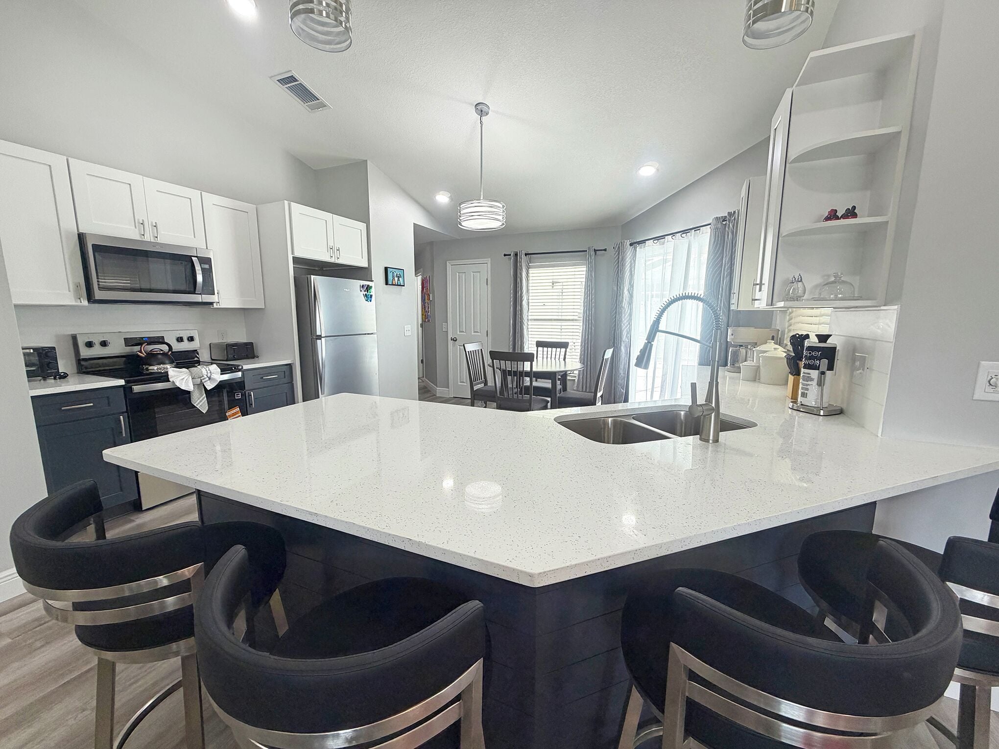 A modern kitchen with a central island, bar stools, stainless steel appliances, and a dining area with a window.