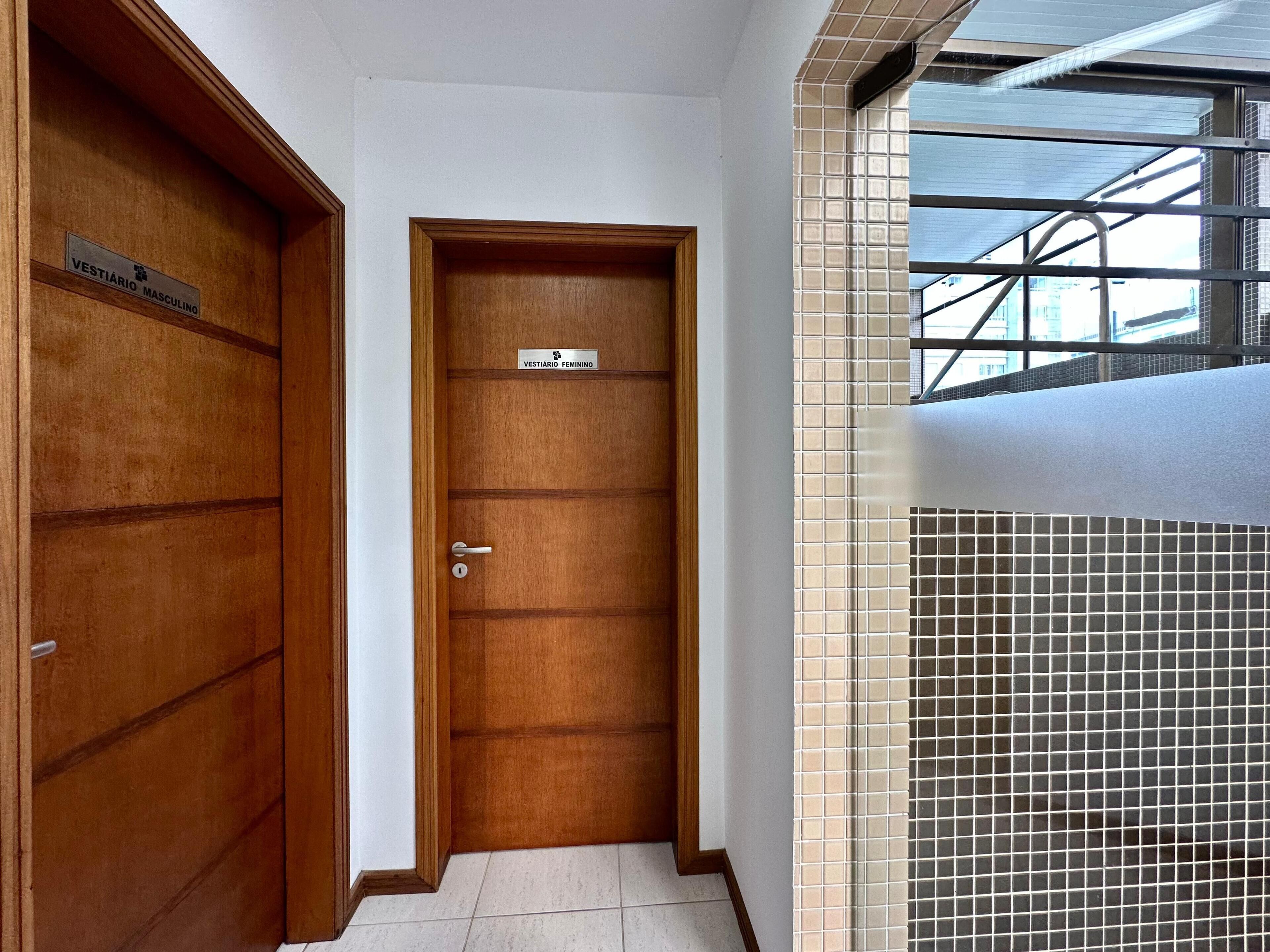 Corredor com duas portas de madeira, uma identificada como 'VESTIÁRIO MASCULINO' e a outra 'VESTIÁRIO FEMININO'.