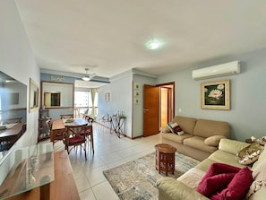 A living room with a sofa, a dining table, and chairs. There is a small wooden stool and a glass table.