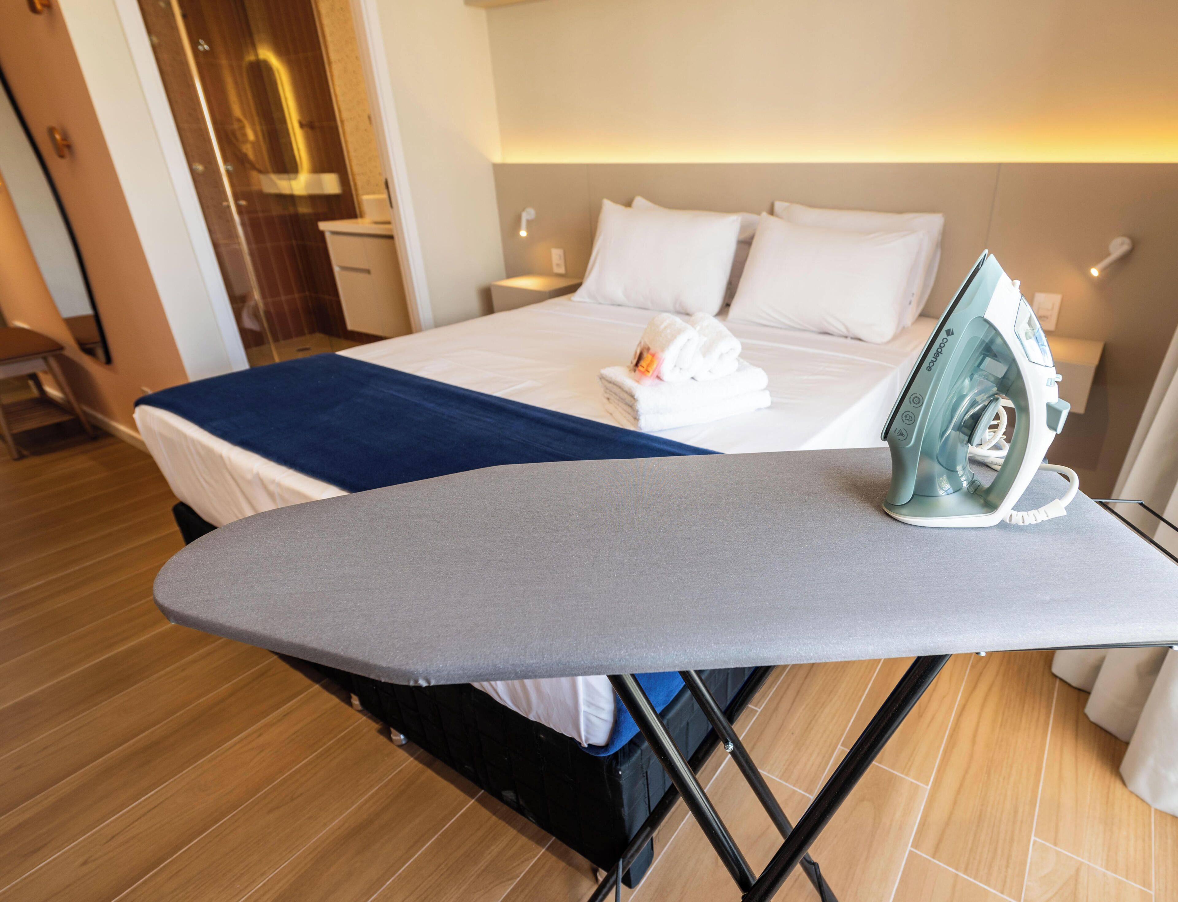 A hotel room with a bed, a bedside table, a suitcase, and an ironing board with an iron.