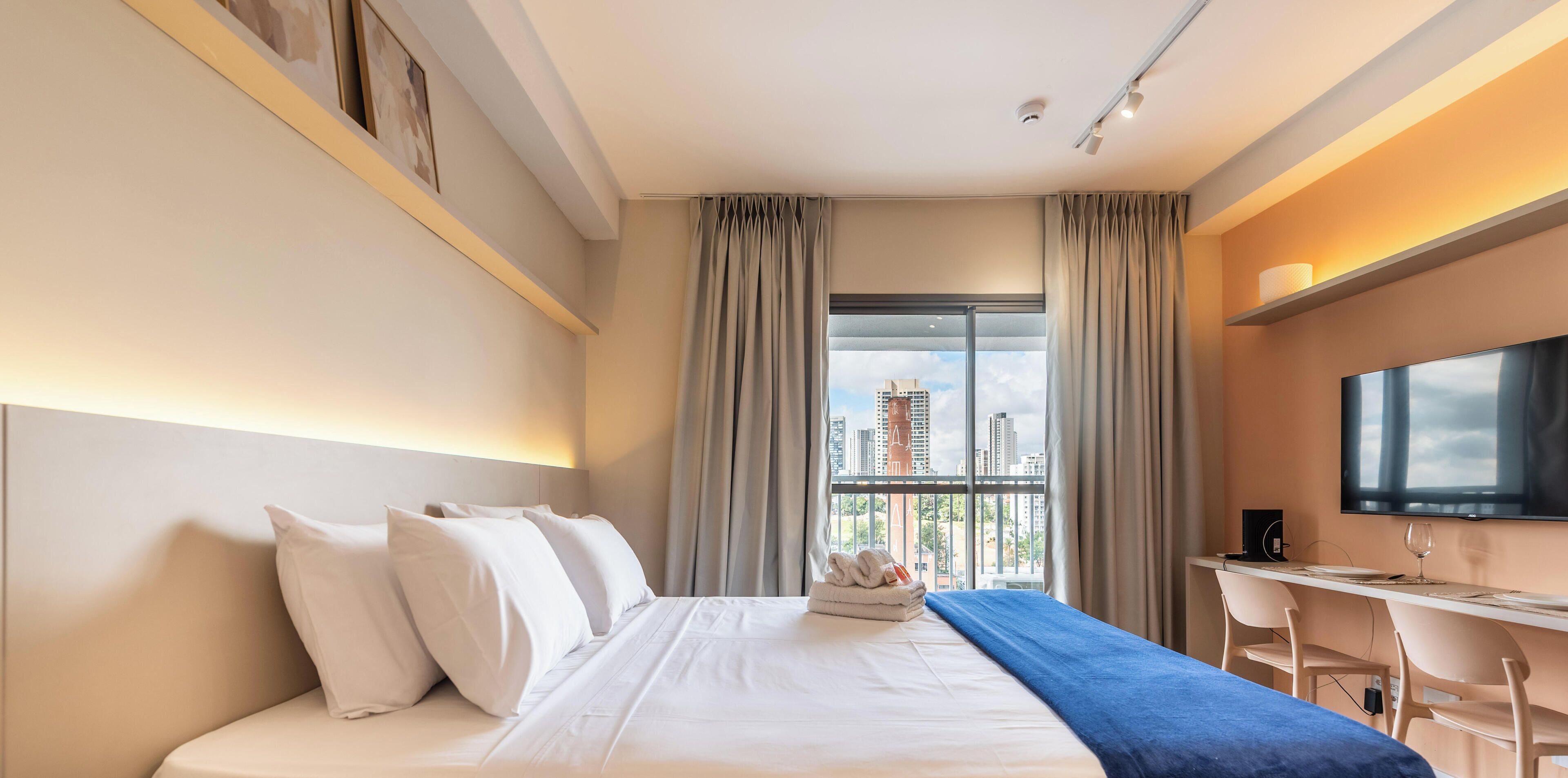 Ein Hotelzimmer mit Bett, Schreibtisch, Stuhl, Fernseher und einem Fenster mit Blick auf die Stadt.