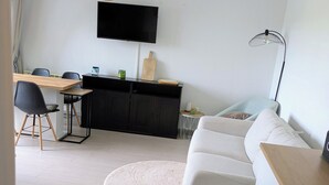 Una sala de estar moderna con un televisor de pantalla plana, una mesa de comedor con sillas, un sofá, una pequeña mesita auxiliar y una lámpara.