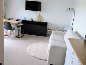 Ett modernt vardagsrum med en platt-TV, ett matbord med stolar, en soffa, ett litet sidobord och en lampa.