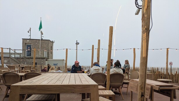 Una terraza de madera con mesas y sillas, un edificio de piedra con una bandera, y vista al mar.