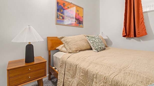 A bedroom with a bed, a nightstand, a lamp, a painting, and a curtain. - New 2BR Skywalk Apt+Downtown Indy+Sleeps 7 (Indianapolis)