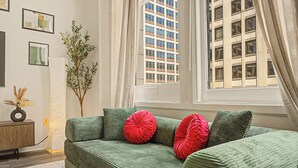 A green sofa with red cushions, a small wooden side table with a vase, and a tall plant. - New 2BR Skywalk Apt+Downtown Indy+Sleeps 7 (Indianapolis)