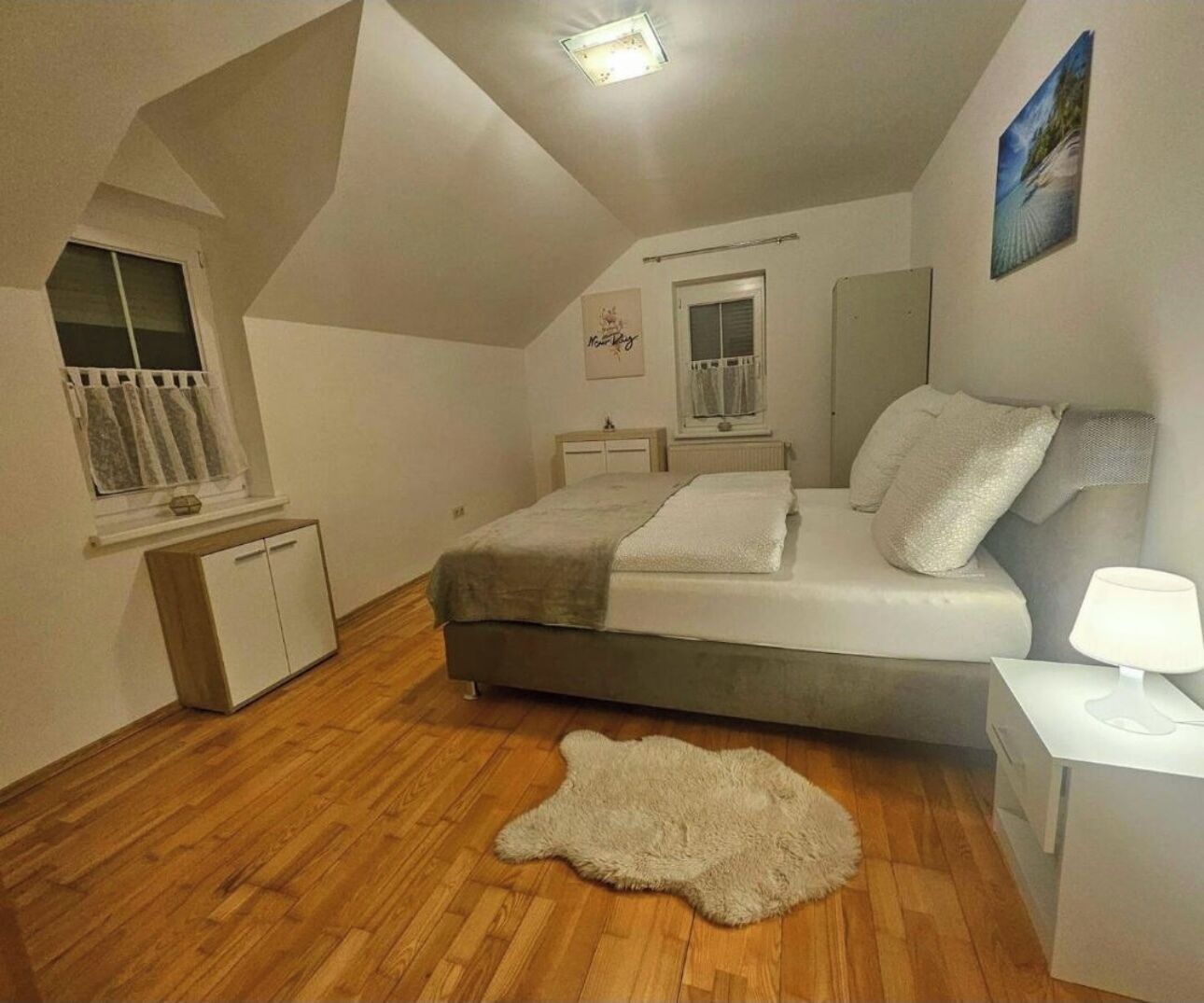 A bedroom with a bed, a nightstand, a lamp, a window with curtains, a wardrobe, and a rug.