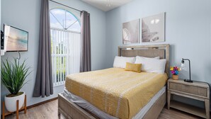 A bedroom with a bed, a nightstand, a TV, a potted plant, and a window with curtains. - 3BR Home with Private Pool Minutes from Disney (Kissimmee)