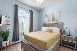 A bedroom with a bed, a nightstand, a TV, a potted plant, and a window with curtains. - 3BR Home with Private Pool Minutes from Disney (Kissimmee)