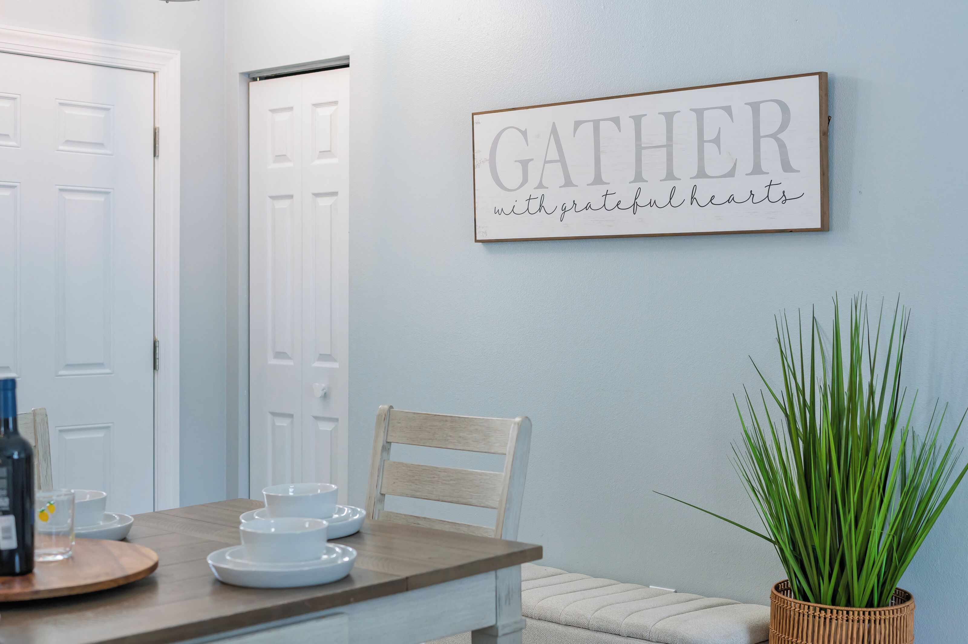A dining area with a table set for four, a wall-mounted sign that reads 'GATHER with grateful hearts,' and a potted plant on the right.