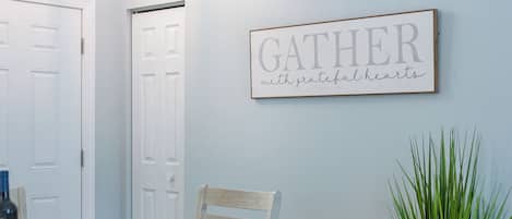 A dining area with a table set for four, a wall-mounted sign that reads 'GATHER with grateful hearts,' and a potted plant on the right.