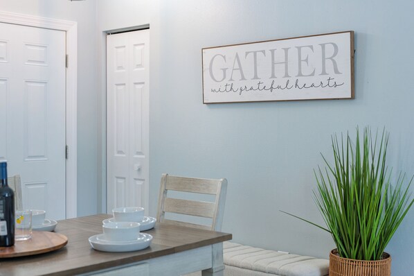 Ein Essbereich mit einem Tisch für vier Personen, einem an der Wand befestigten Schild mit der Aufschrift 'GATHER with grateful hearts' und einem Blumentopf auf der rechten Seite.