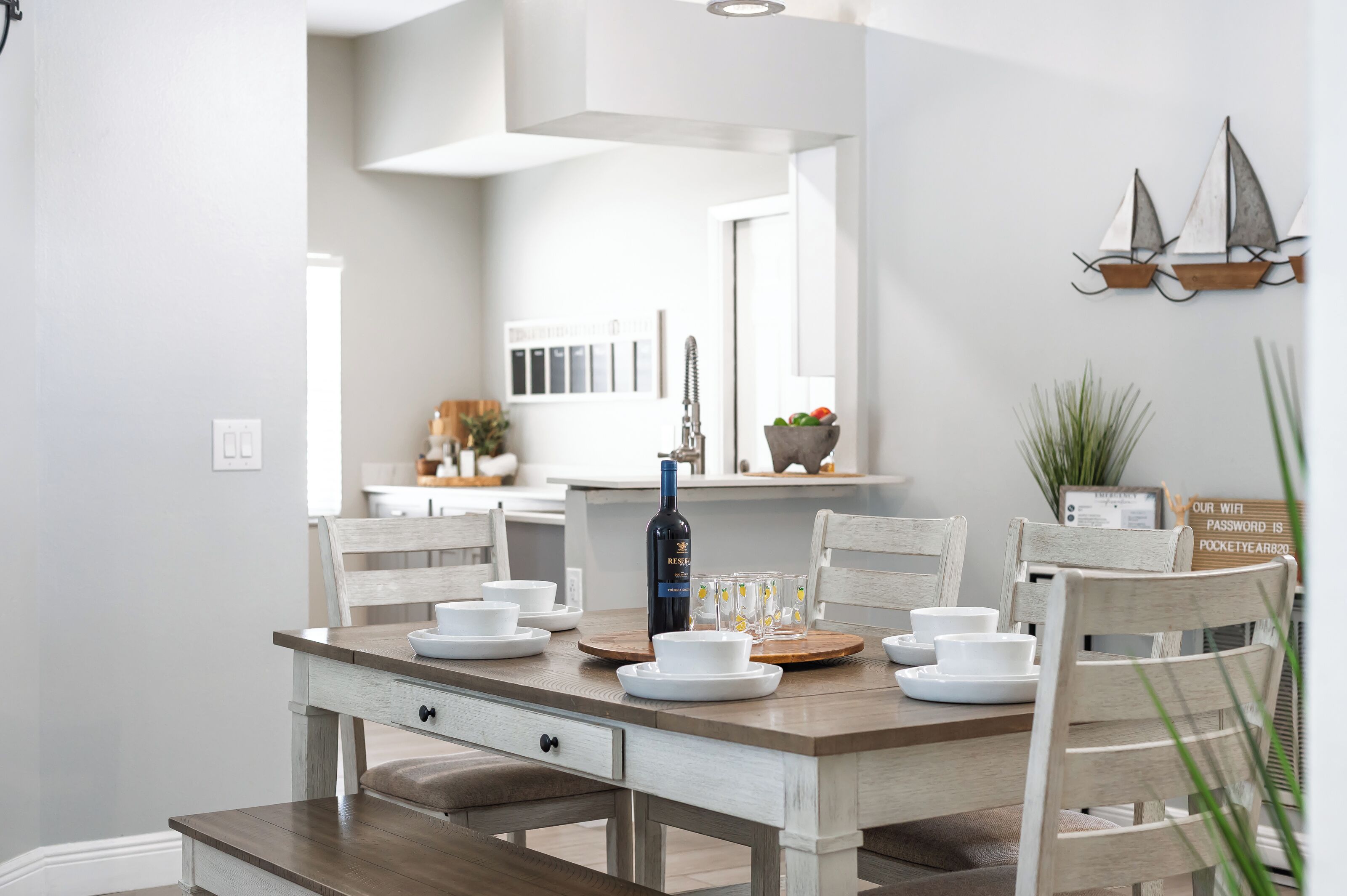 A dining area with a wooden table set for four, white chairs, and a bottle of wine.