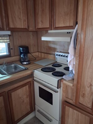 A compact kitchen with wooden cabinets, a stove, a sink, and a coffee maker. - Beautiful 1-bedroom mobile home with AC in Jensen Beach (Jensen Beach)
