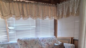 A room with a floral-patterned sofa, a pair of shoes on the floor, and a window with blinds. - Beautiful 1-bedroom mobile home with AC in Jensen Beach (Jensen Beach)
