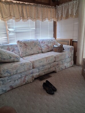 A room with a floral-patterned sofa, a pair of shoes on the floor, and a window with blinds. - Beautiful 1-bedroom mobile home with AC in Jensen Beach (Jensen Beach)