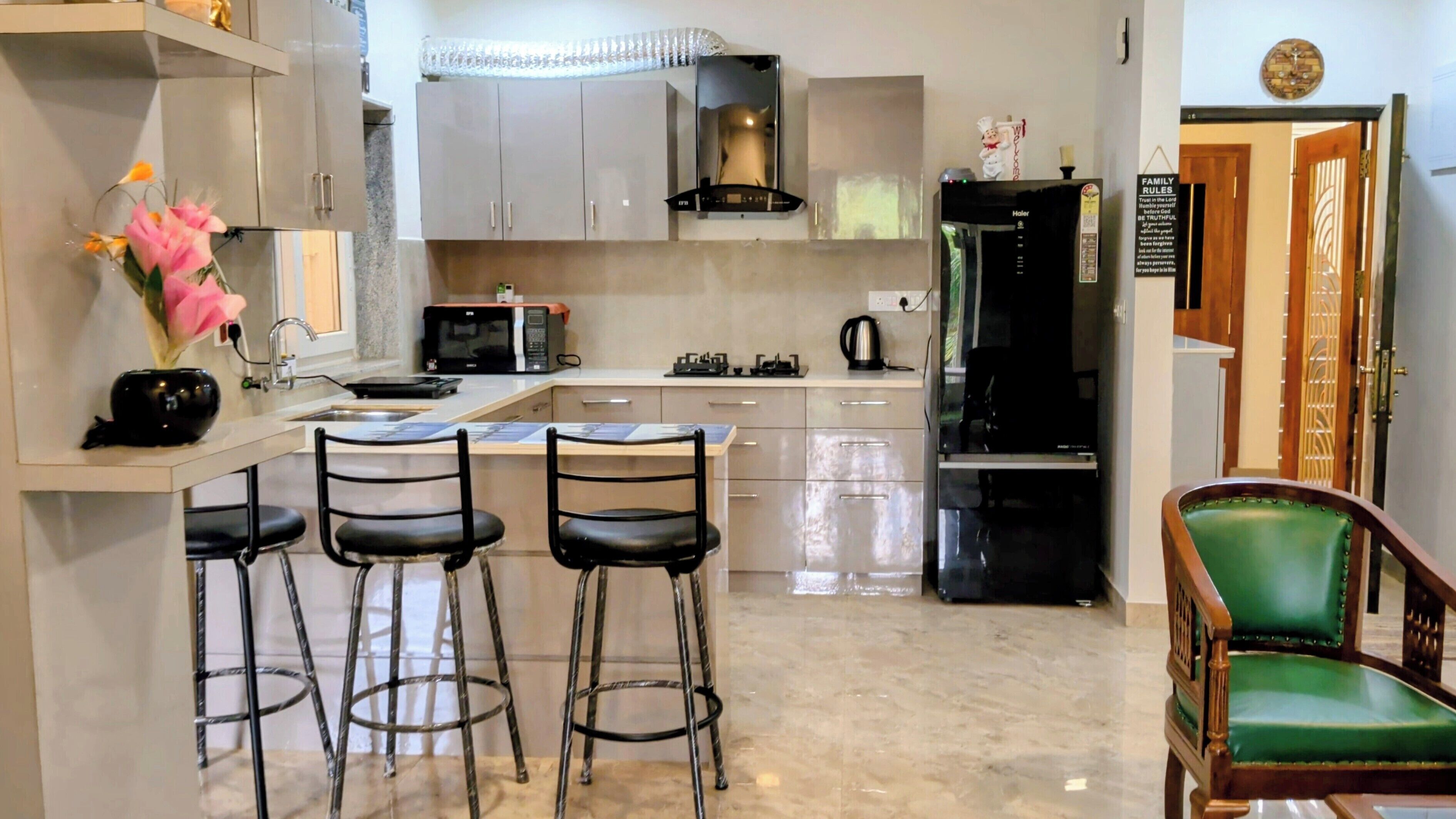 A kitchen with a microwave, stove, and refrigerator. There are three bar stools and a green chair.
