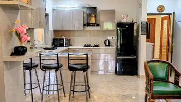 A kitchen with a microwave, stove, and refrigerator. There are three bar stools and a green chair.