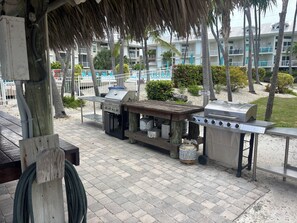 Una estación de parrilla al aire libre con mesa de madera, una parrilla a gas y una parrilla de carbón, rodeada por un patio empedrado con una palmera y una piscina al fondo.