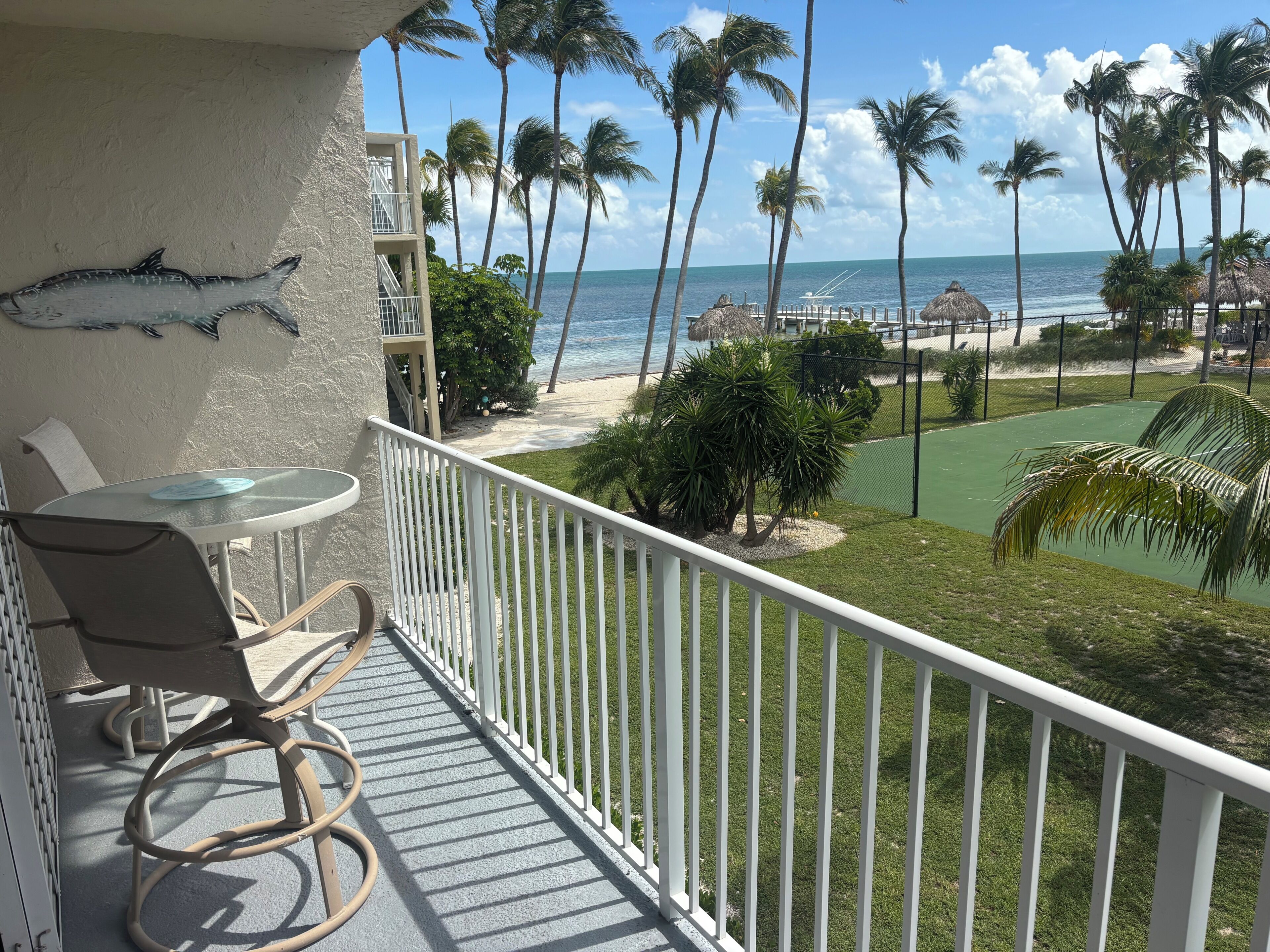 A balcony with a table and chairs, overlooking a beach with palm trees and a clear sky.