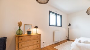 A bedroom with a wooden dresser, a framed picture, a lamp, a radiator, a window, and a bed with a white blanket. - Architect-designed house 800m from the sea (Paimpol)