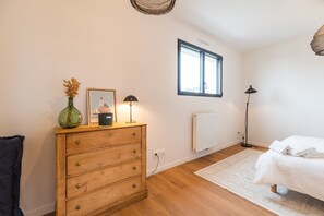 A bedroom with a wooden dresser, a framed picture, a lamp, a radiator, a window, and a bed with a white blanket. - Architect-designed house 800m from the sea (Paimpol)