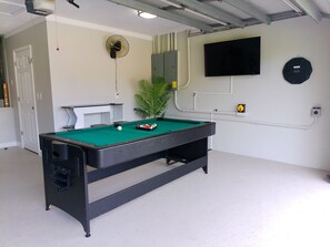 Ein Billardtisch in einem Raum mit Tür, Ventilator, Pflanze und einem angebrachten Fernseher.
