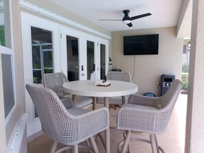Eine Terrassenfläche mit einem runden Tisch, vier Korbstühlen, einem Deckenventilator, einem an der Wand montierten Fernseher und einem Terrassenheizer.