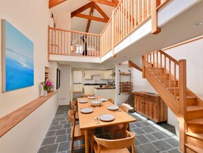 A spacious dining area with a high ceiling and exposed wooden beams, a kitchen in the background, and a staircase leading to an upper level. - Village Home With Sea Views (Truro)