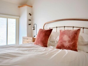 A neatly made bed with white bedding and two pink pillows, a bedside table with a lamp, and a window providing natural light. - Village Home With Sea Views (Truro)