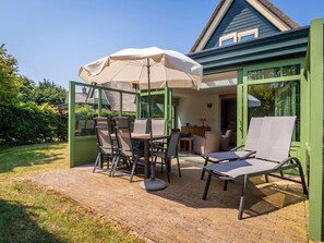 Eine Terrasse im Freien mit Tisch und Stühlen, einem beschatteten Sonnenschirm und Blick auf ein Haus mit grünen Türen und Fenstern.