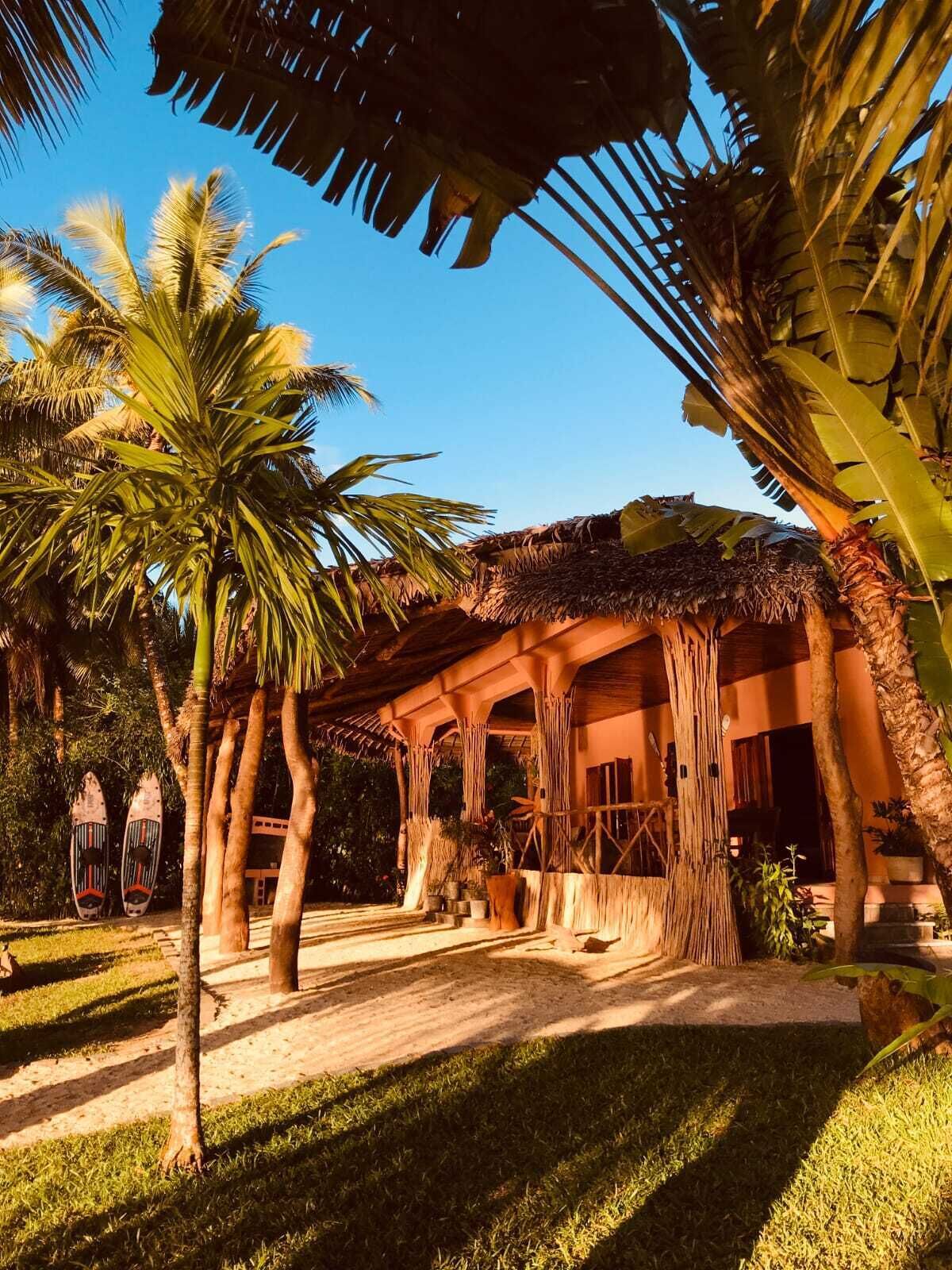 A tropical resort with a thatched-roof structure, palm trees, and a clear blue sky.