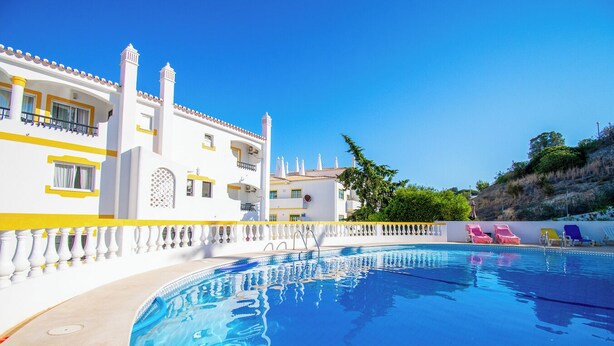 A swimming pool with a white and yellow building in the background, a white fence, and a hillside with trees. - 2 mins walk to Carvoeiro beach, Amazing Pool, Aircon, newly renovated, Fast WiFi (Carvoeiro)