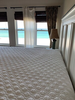 Uma cama com cobertura branca texturizada, vista para o mar através de uma janela e uma luminária em uma mesa de cabeceira.