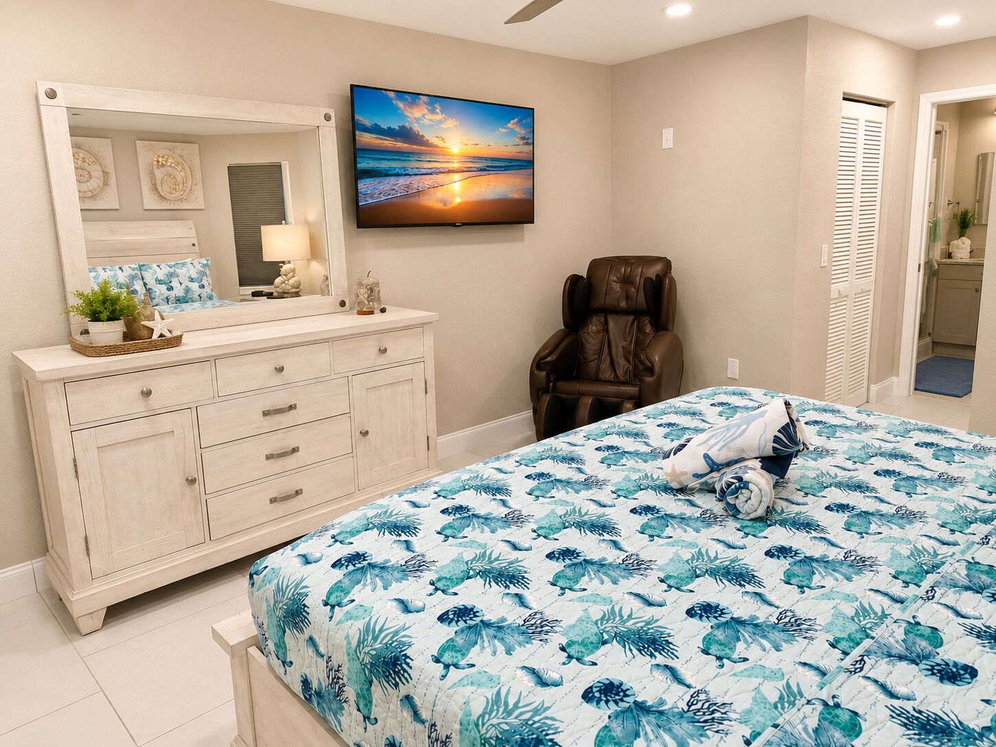 Une chambre à coucher avec un lit, un fauteuil de massage, une télévision et une salle de bain visible à travers une porte ouverte.