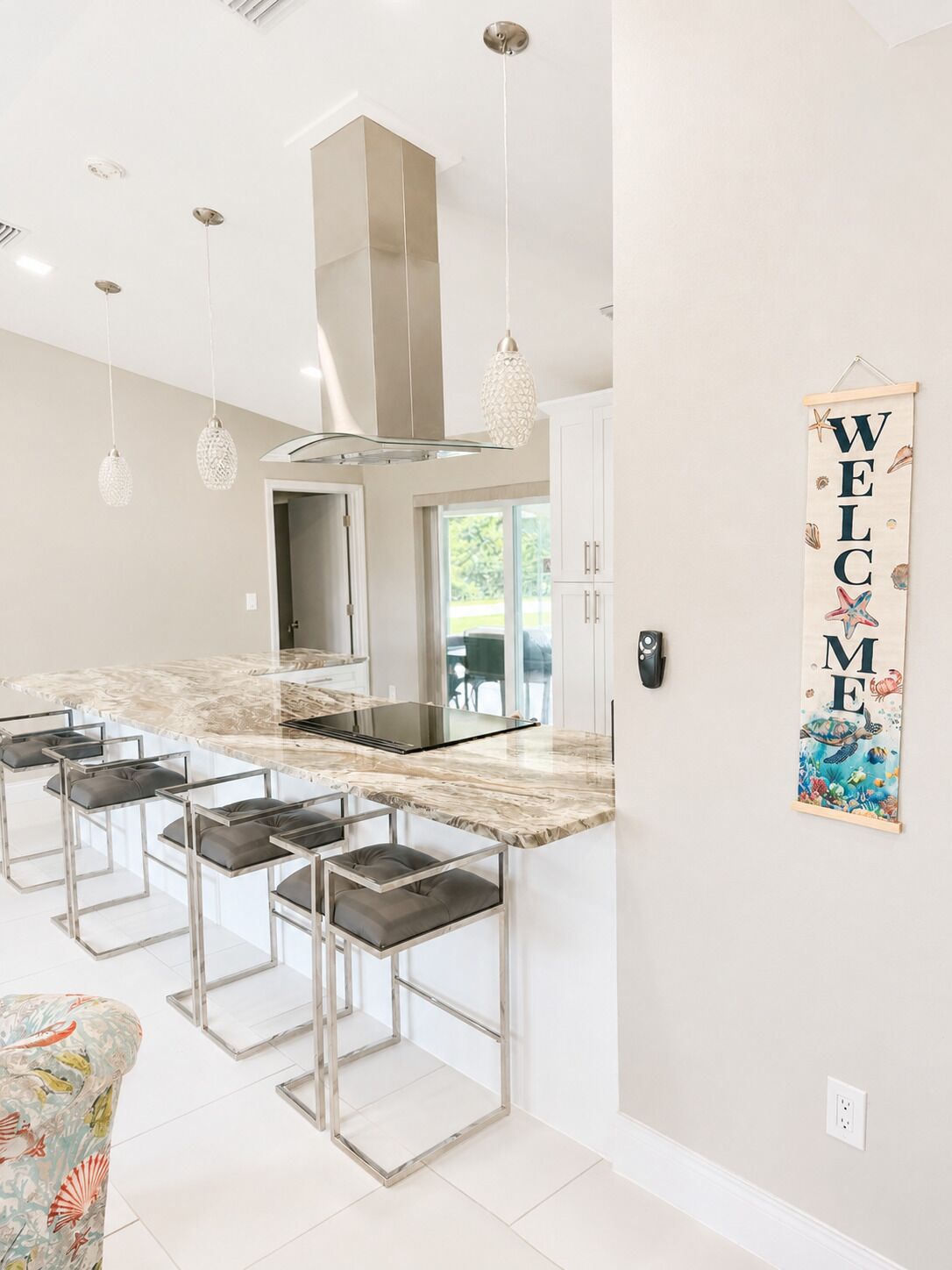 A modern kitchen with a marble countertop, bar stools, pendant lights, and a welcome sign.
