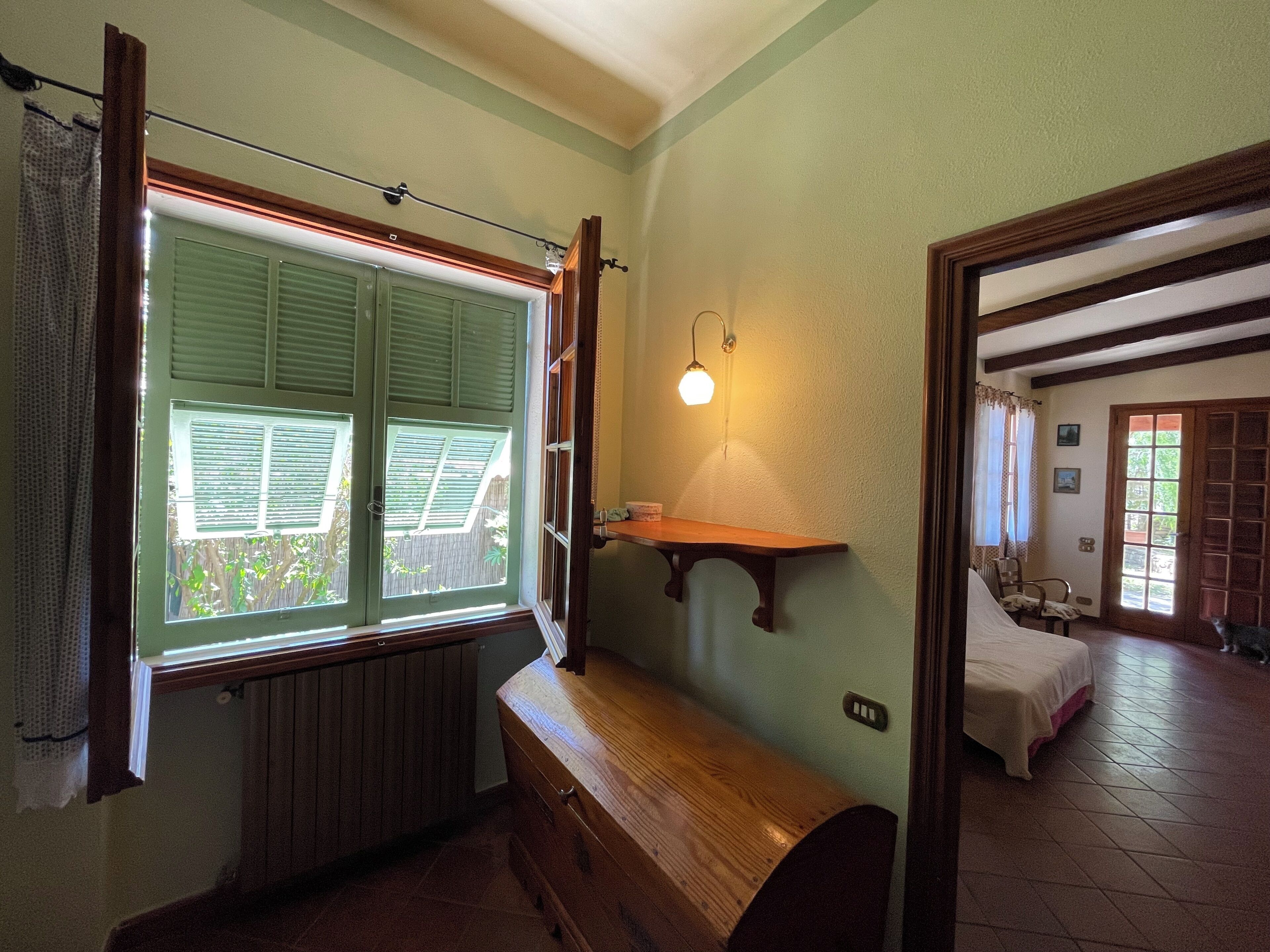 A room with a wooden bench, a wall-mounted shelf, a window with shutters, and a door leading to another room.