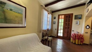 Ein Zimmer mit einer gepolsterten Couch, einem Esstisch mit Tischdecke, einem Stuhl, einer Tür mit Glasfenstern und einem gerahmten Bild an der Wand.