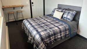 Een slaapkamer met een bed, een bureau met twee krukken, een deur en een lichtschakelaar aan de muur.