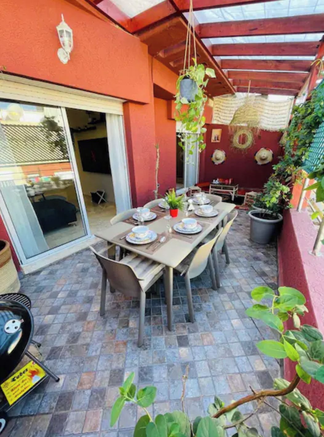Un espace extérieur aménagé pour prendre les repas, avec une table dressée pour deux personnes, entourée de plantes en pot et bordée d’un mur rouge.